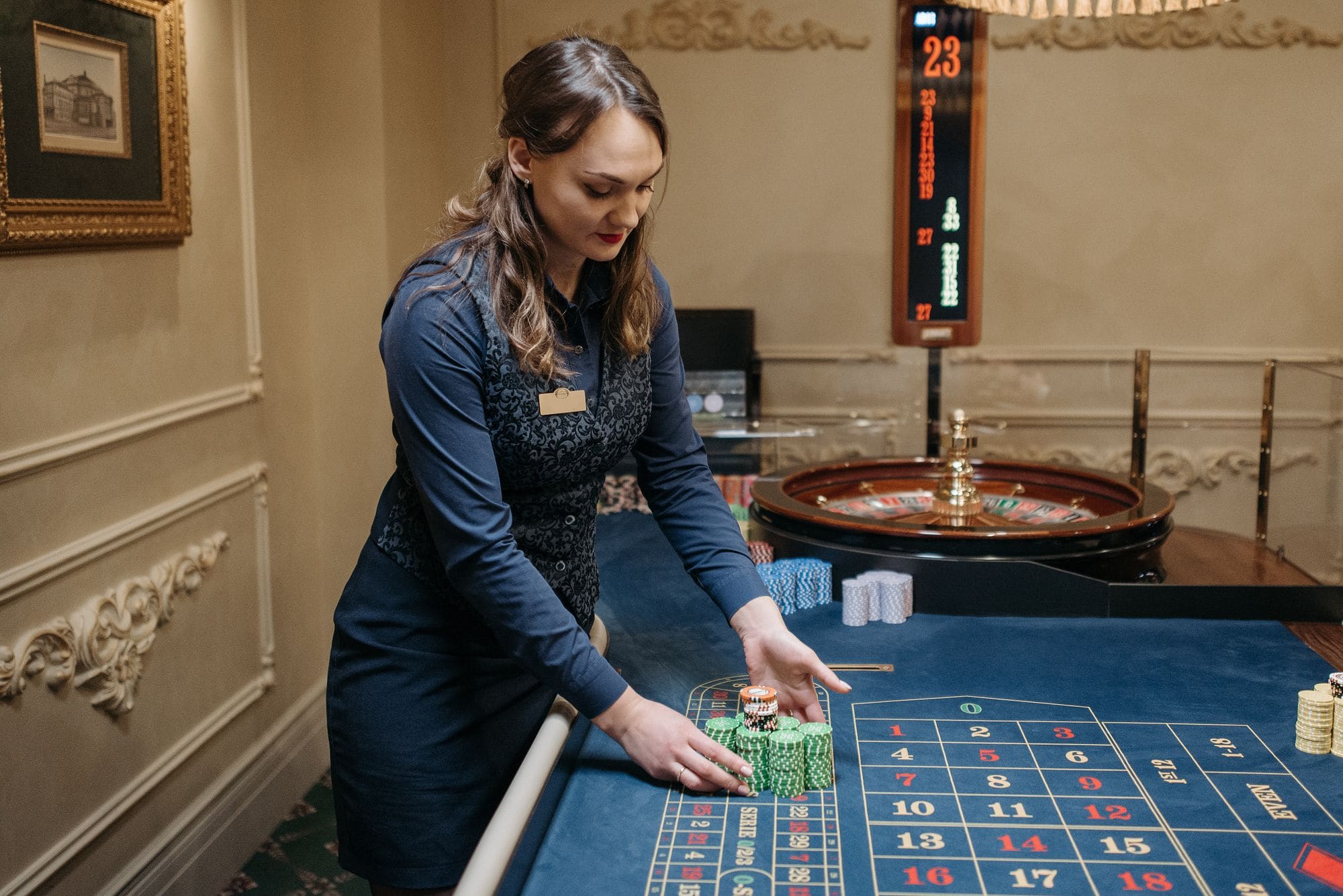A croupier in a dark waistcoat placing chips on the layout of a roulette table in a chandeliered gaming room
