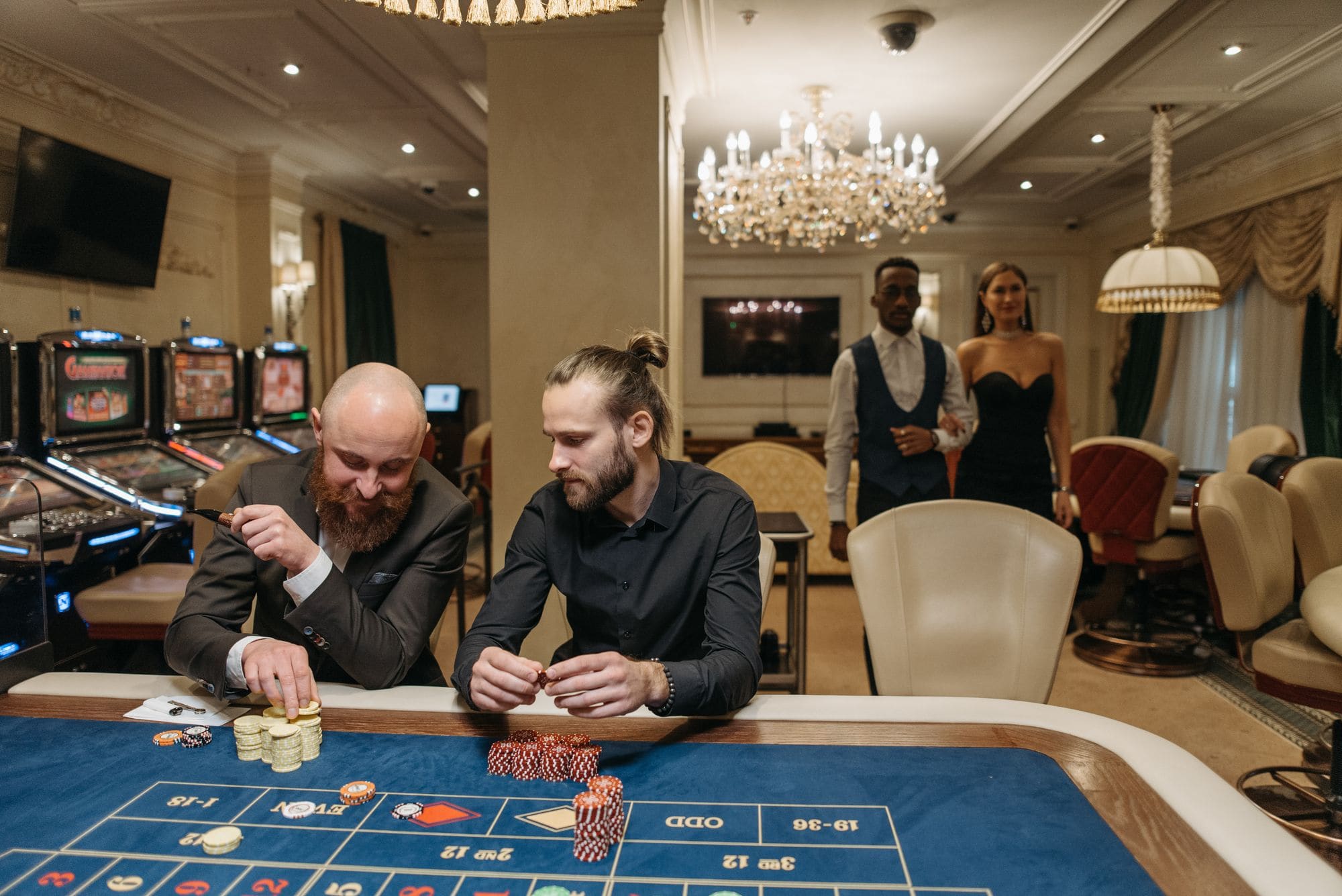 Players seated at a roulette table in an elegant casino lounge with a crystal chandelier overhead